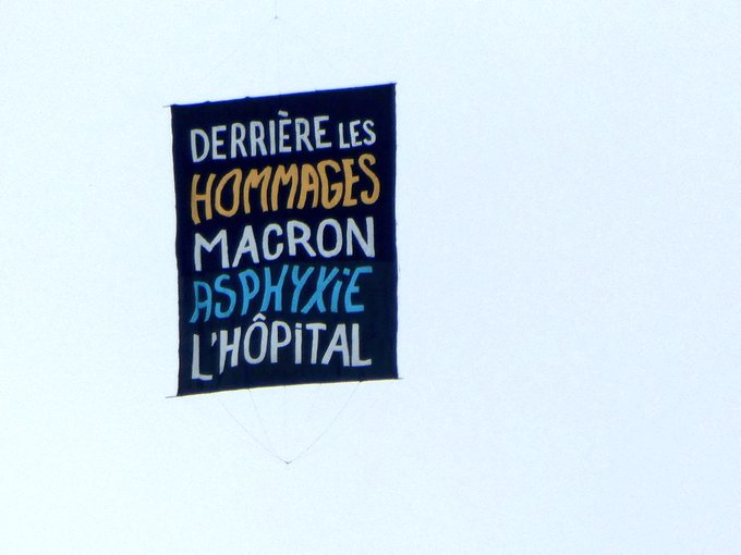 Pendant la Marseillaise, des ballons et une banderole s’envolent : “L’économie nous coûte la vie” et “derrière les hommages Macron asphyxie l’hôpital”.